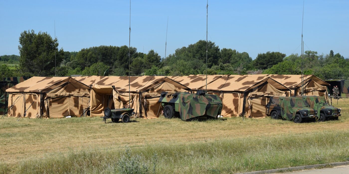 Vue aérienne d'un poste de commandement de brigade : abris I-4S raccordés en ligne, véhicules et conteneurs déployés en lisière de bois